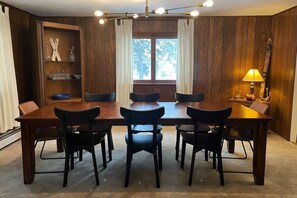 A dining area with a wooden table and chairs, a window with curtains, and a chandelier. - The Rose Lodge: 8 Bedrooms, 7 Baths, Private Hot Tub and Game Room (Killington)