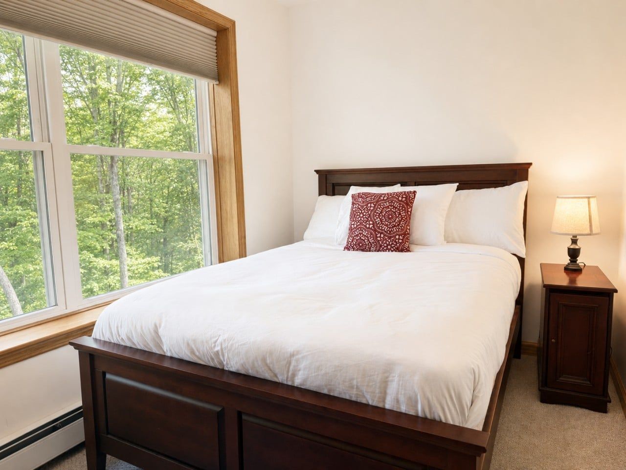 A bedroom with a large bed, a nightstand, a lamp, and a window with a view of trees.