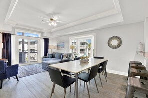 A modern living room with a dining area, a ceiling fan, and a view through the glass doors. - Ocean-view 3BR condo with pool accces (Tybee Island)
