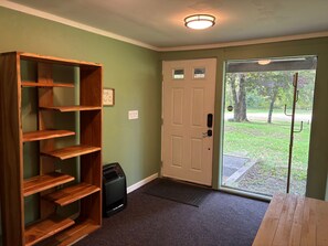 A room with a wooden shelving unit, a door with a peephole, and a view of a grassy area through a glass door. - Riverview Retreat | beach, float, ski (Campton)
