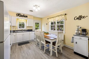 A kitchen with white cabinets, a wooden dining table, and chairs. There is a window with a floral curtain, and a coffee machine on the counter. - 5519 Moffitt's, Sound Side, Community Pool/Tennis (Kill Devil Hills)