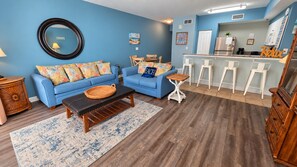 A living room with a blue sofa set, a coffee table, a wooden side table, and a kitchen area with a bar stool. - Shores of Panama 927 - 1 Bedroom+Bunks w/Sunset View (Panama City Beach)