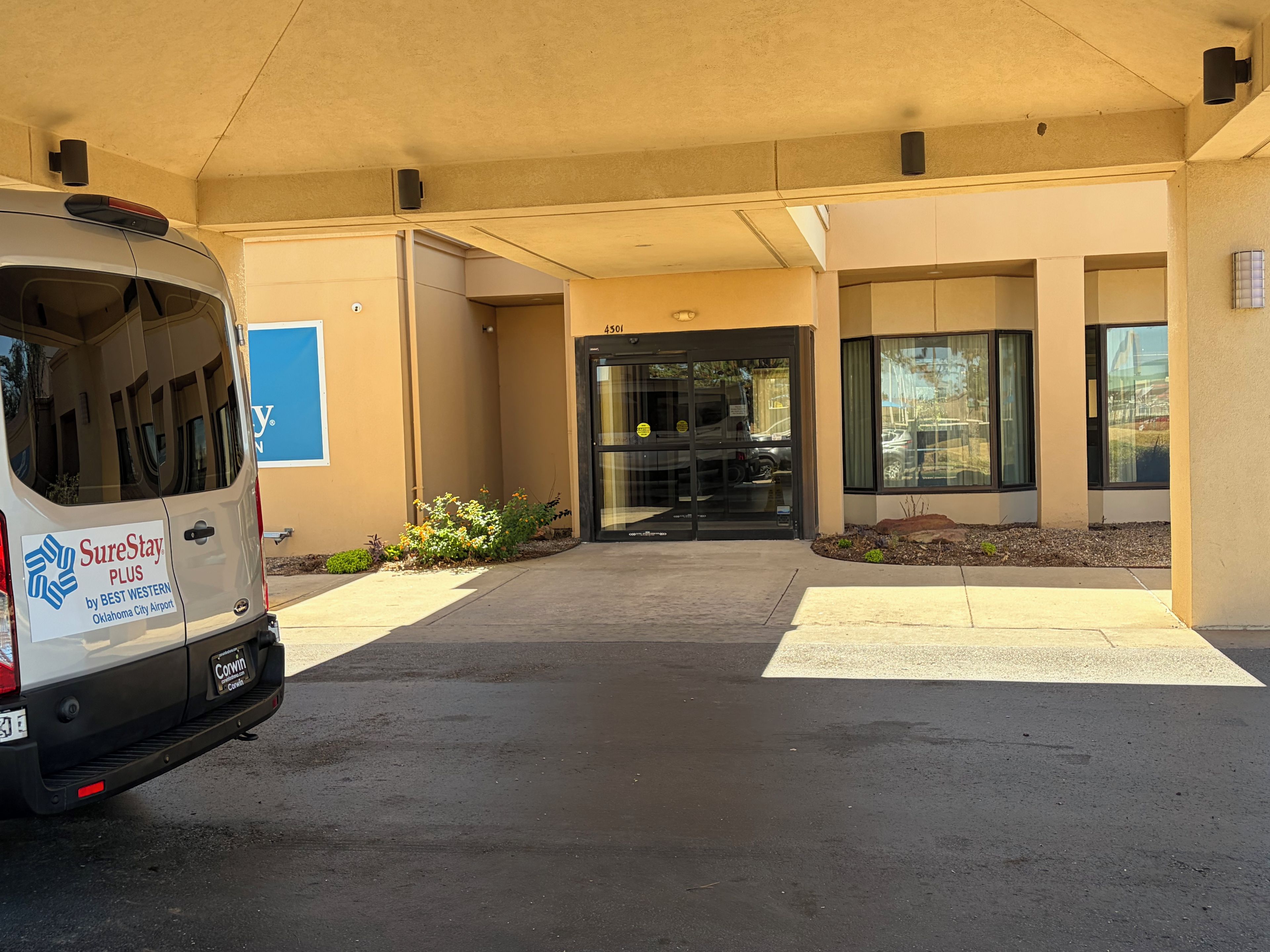 Un área de estacionamiento con una camioneta blanca estacionada frente a un edificio con entrada por puerta de vidrio.