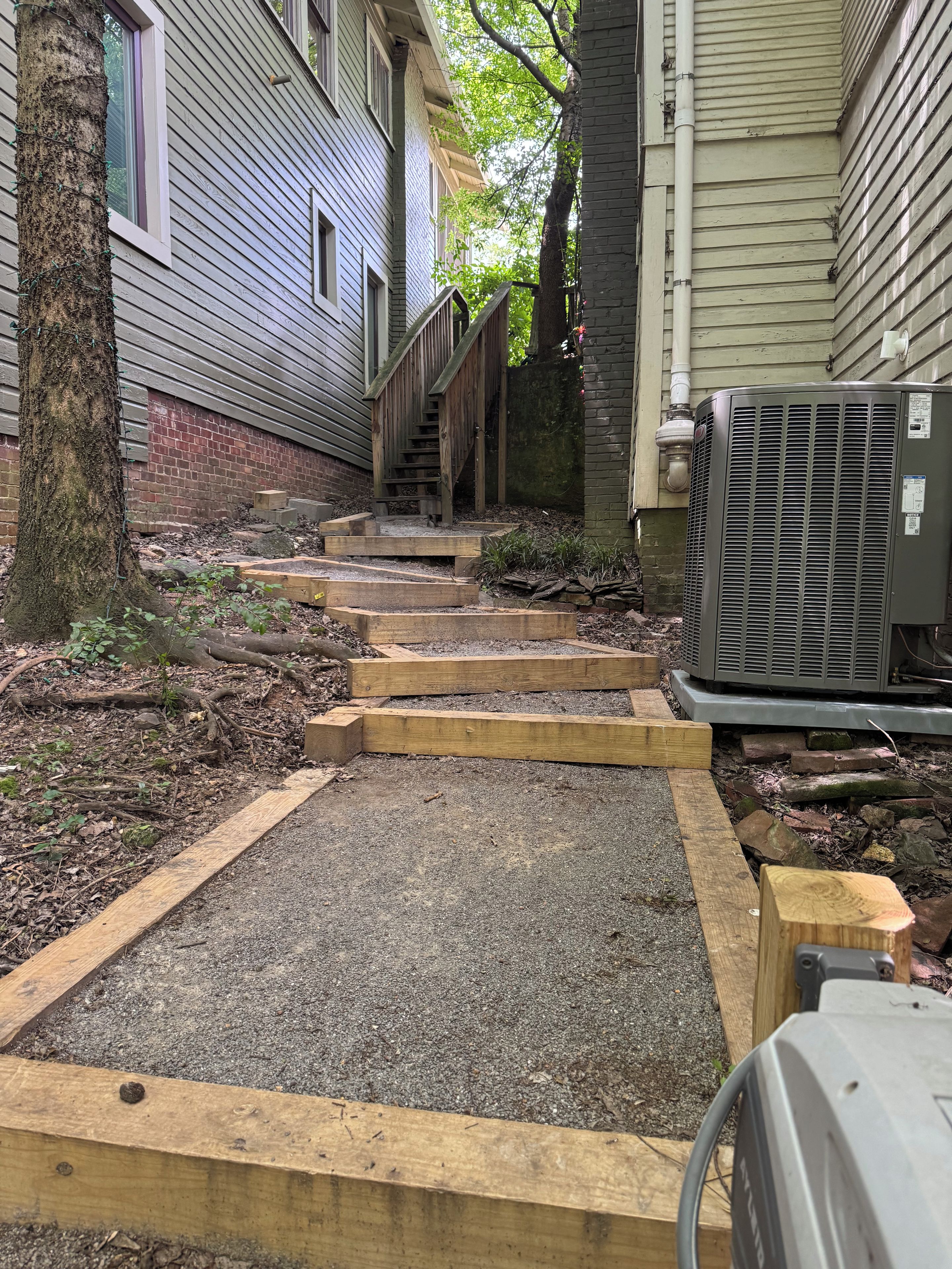 Un sendero lleva a una escalera de madera, con un muro de ladrillo y una unidad de aire acondicionado a la derecha.