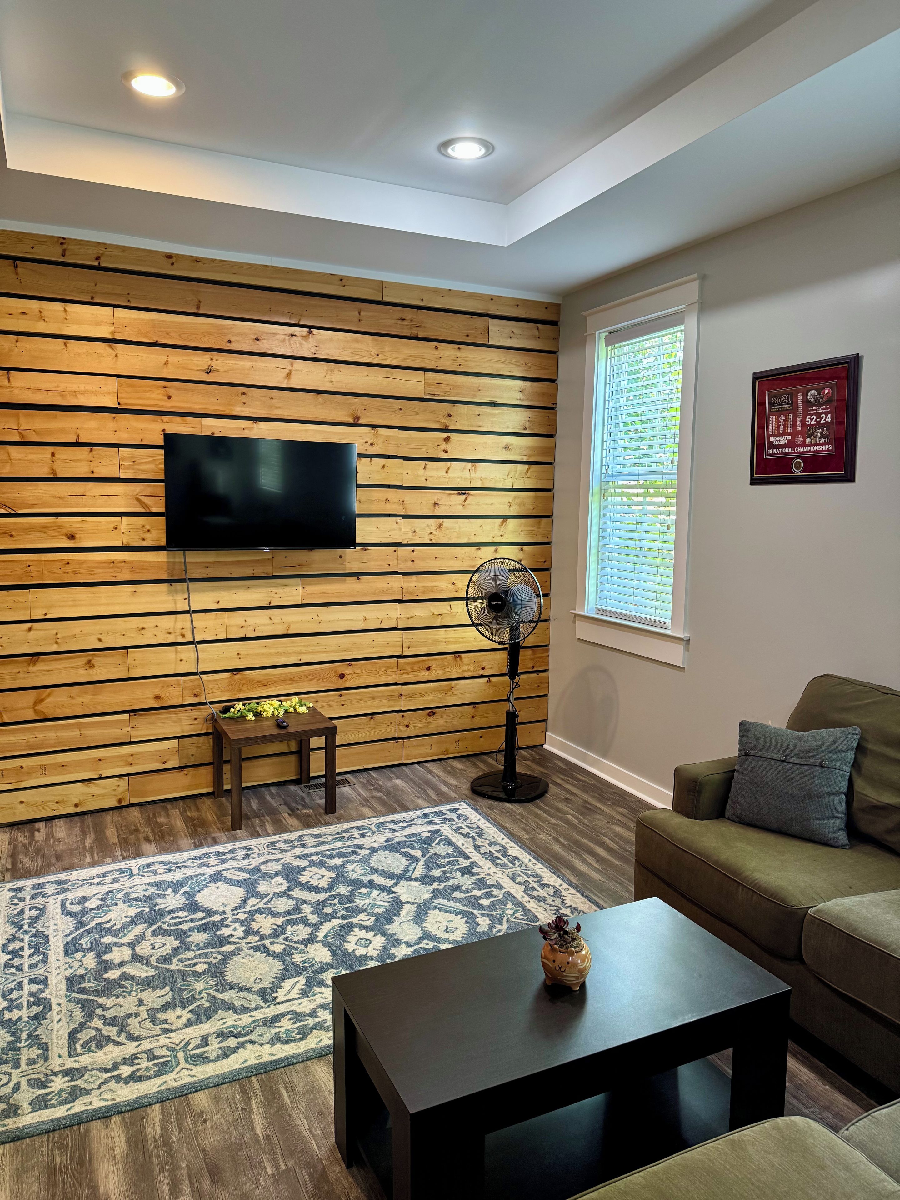 Una sala de estar con un panel de pared de madera, un televisor de pantalla plana, un ventilador de pie, un tapiz estampado, una mesa de centro oscura y un sofá verde.