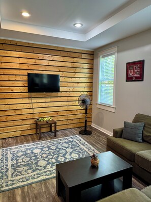 Un salon avec un mur lambrissé en bois, un téléviseur à écran plat, un ventilateur sur pied, un tapis à motifs, une table basse foncée et un canapé vert.