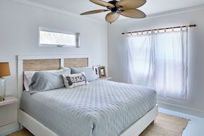 A bedroom with a bed, a ceiling fan, a window with sheer curtains, and a bedside table with a lamp. - Bohemia Cosmo - A Beachfront Bungalow, Pool & Daybed (Governor's Harbour)