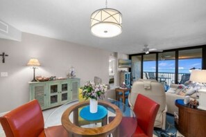 A room with a round table, chairs, a cabinet, a lamp, and a view of the outdoors. - Sunset 304-Charming Beachfront Condo with Gulf Views (Fort Myers Beach)