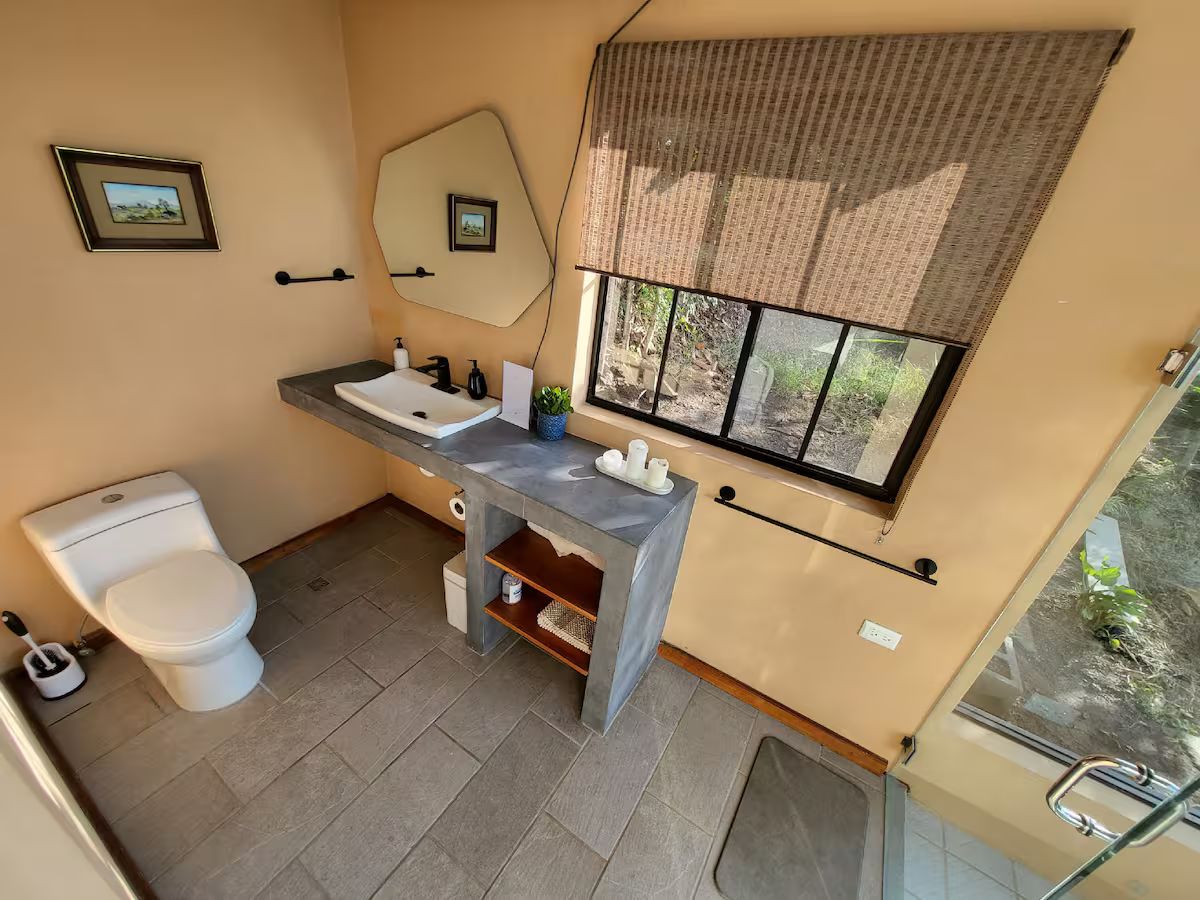 A bathroom with a toilet, sink, mirror, and window.