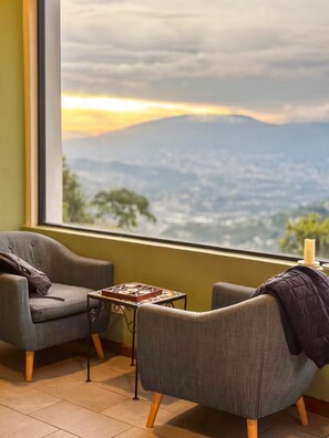A room with two grey armchairs, a small table with a board game, and a view of a mountainous landscape through a large window. - The Monticello Cave (Los Planes de Renderos)