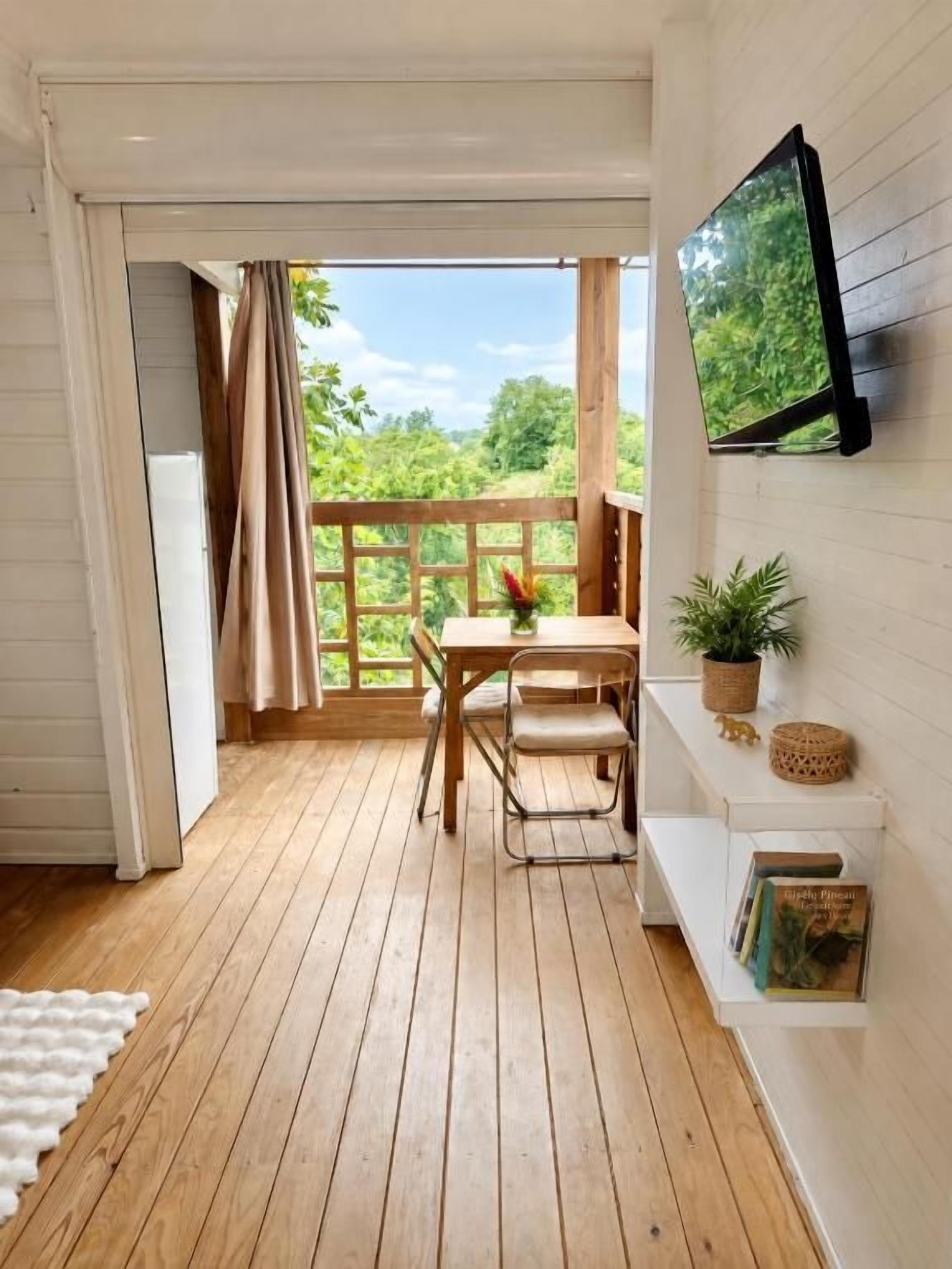 Una habitación con piso de madera, una mesa con sillas, un televisor montado en la pared y un ventanal con cortinas.