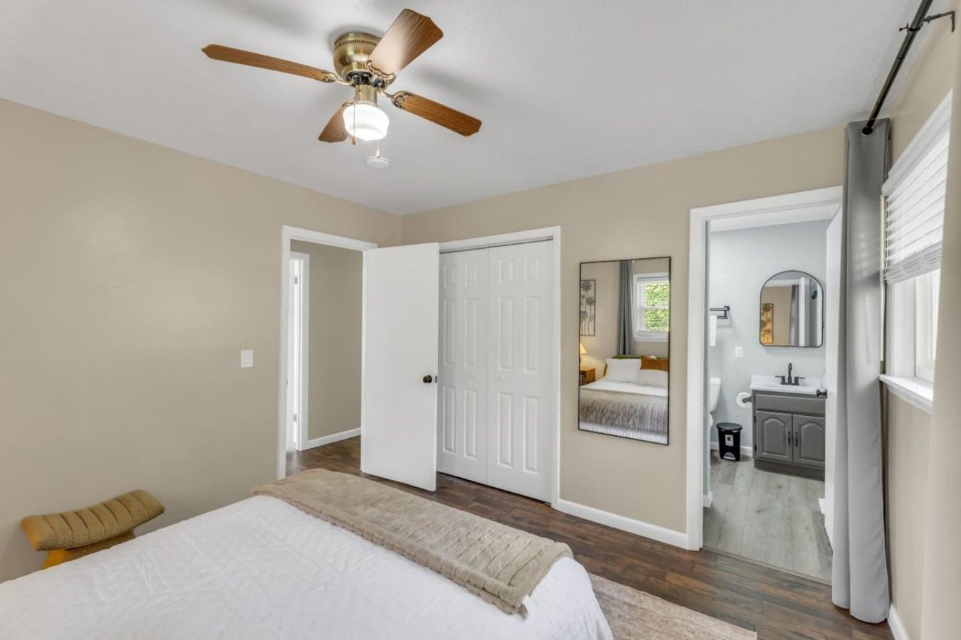 A bedroom with a bed, a ceiling fan, a door leading to a bathroom, and a window with curtains.