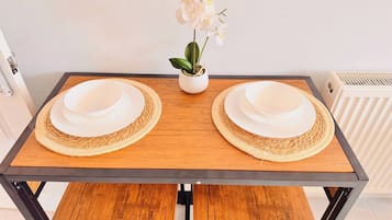 Una mesa rectangular de comedor con dos platos blancos y posavasos tejidos, una orquídea en maceta y un radiador al fondo.