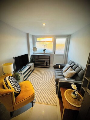 A modern living room with a sofa, a television, a small table with a lamp, and a window with a view. - Shal Inn at Heathrow Free Parking and Ride on Request (Slough)