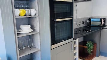 Una cocina compacta con horno empotrado, tostadora y una estantería con cristalería y vajilla.