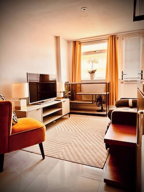 A modern living room with a flat-screen TV, a sofa, a coffee table, and a dining area with a fan. - Shal Inn at Heathrow Free Parking and Ride on Request (Slough)