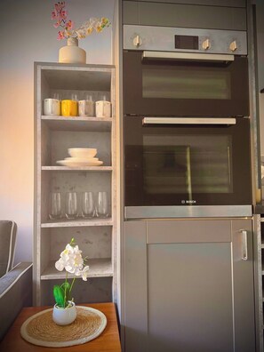 A kitchen with a Bosch oven and a shelf unit containing dishes and glasses. - Shal Inn at Heathrow Free Parking and Ride on Request (Slough)