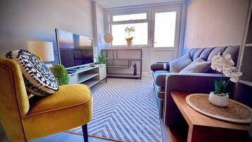 A living room with a sofa, a yellow armchair, a TV, a coffee table, and a window with a view.