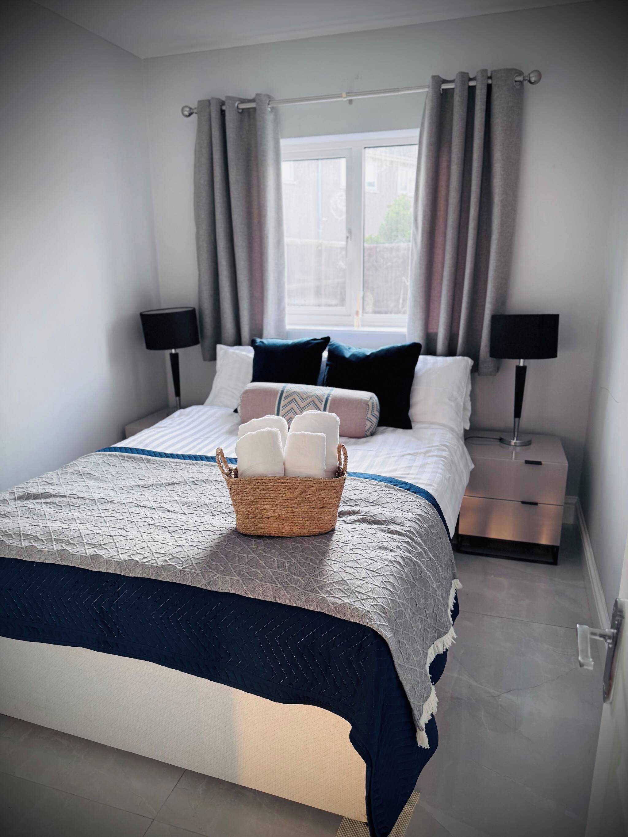 Una cama bien hecha con una cesta de toallas dobladas, dos lámparas de noche y una ventana con cortinas.