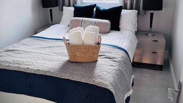 Una cama bien hecha con una cesta de toallas dobladas, dos lámparas de noche y una ventana con cortinas.