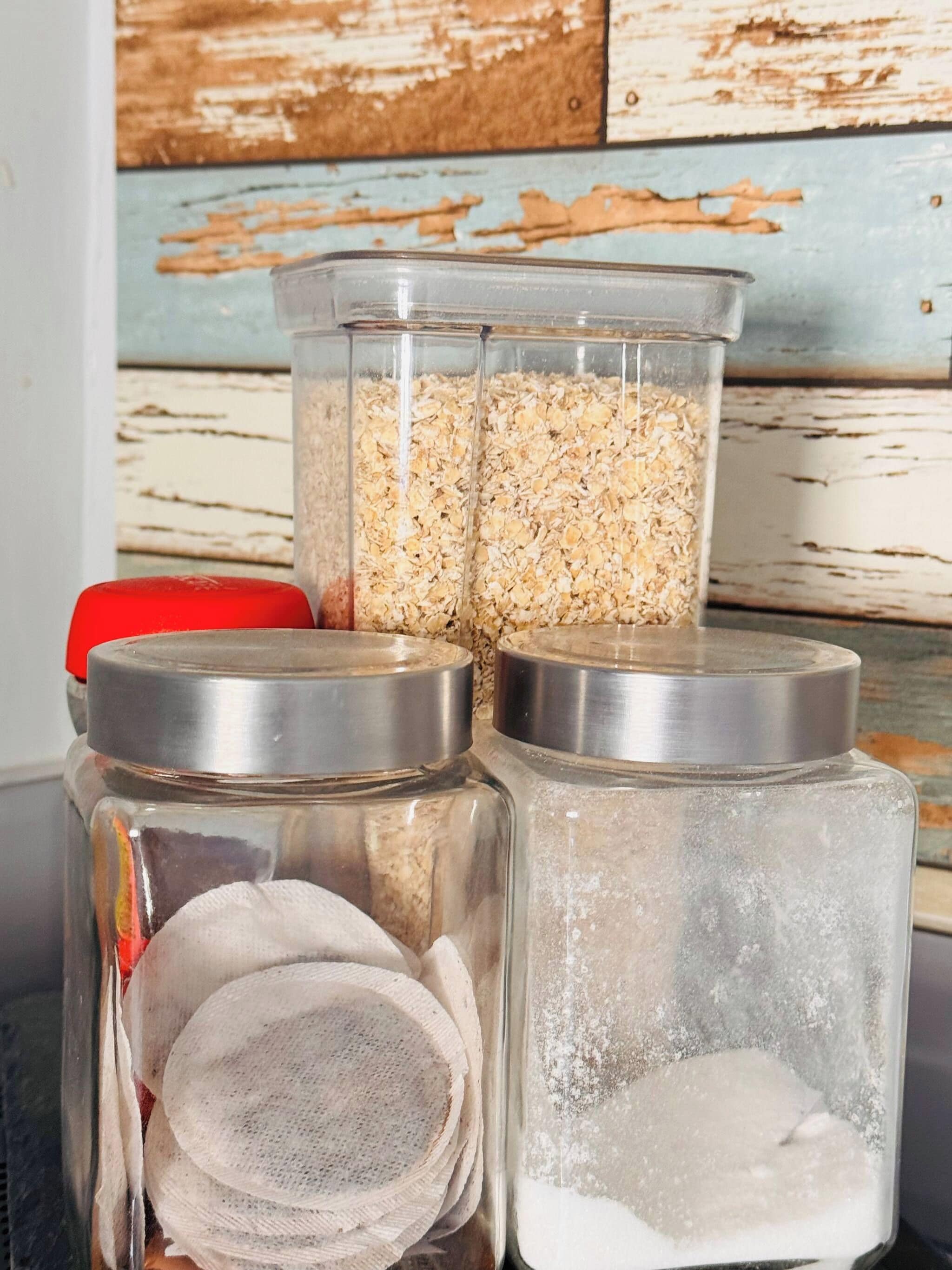 Three glass jars with lids, one containing oats, another with a cloth, and the third with a white powdery substance.