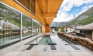 A modern indoor pool with glass walls, wooden ceiling, and a view of a mountainous landscape. - A MODERN ALPINE STYLE APARTMENT WITH  BREATHTAKING DIRECT VIEW OF THE MATTERHORN (Zermatt)