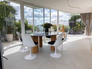 A modern dining area with a glass table surrounded by white chairs, large windows, and a view of greenery. - Le Rooftop Eiffel & La Défense - Welkeys (Neuilly-sur-Seine)