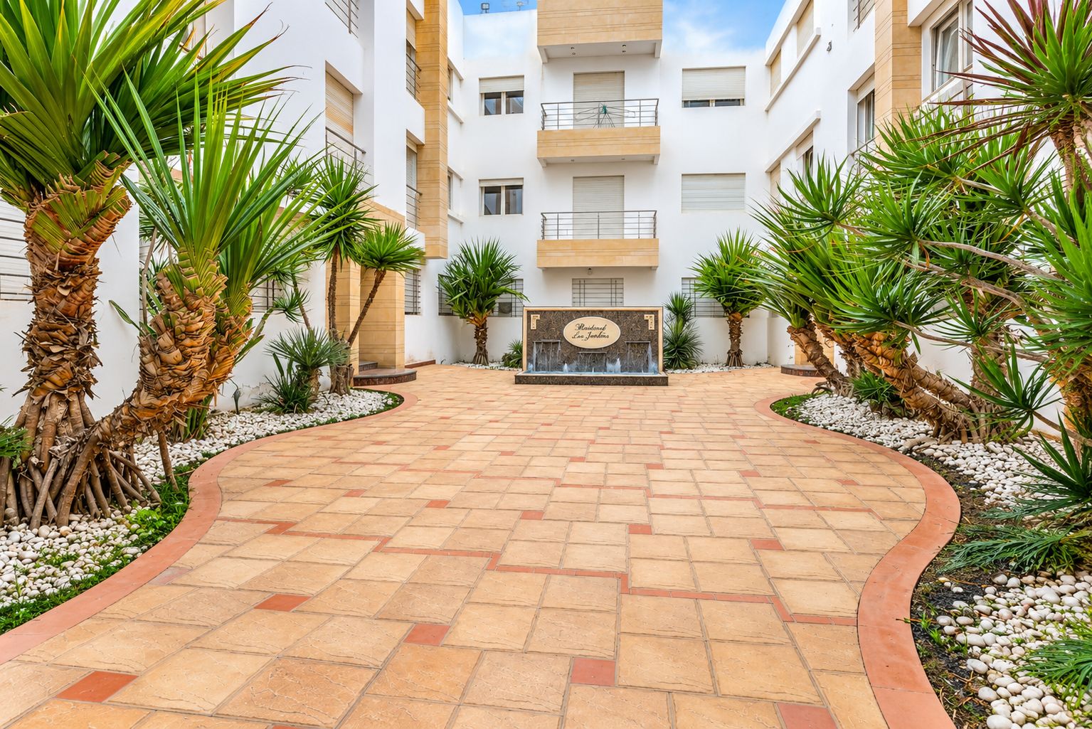 A courtyard with a paved walkway, palm trees, and a building with balconies.