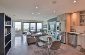 A modern kitchen with stainless steel appliances, a dining area with a glass table, and a view of the sea through large windows. - The Beachcomber Suite (Victoria)