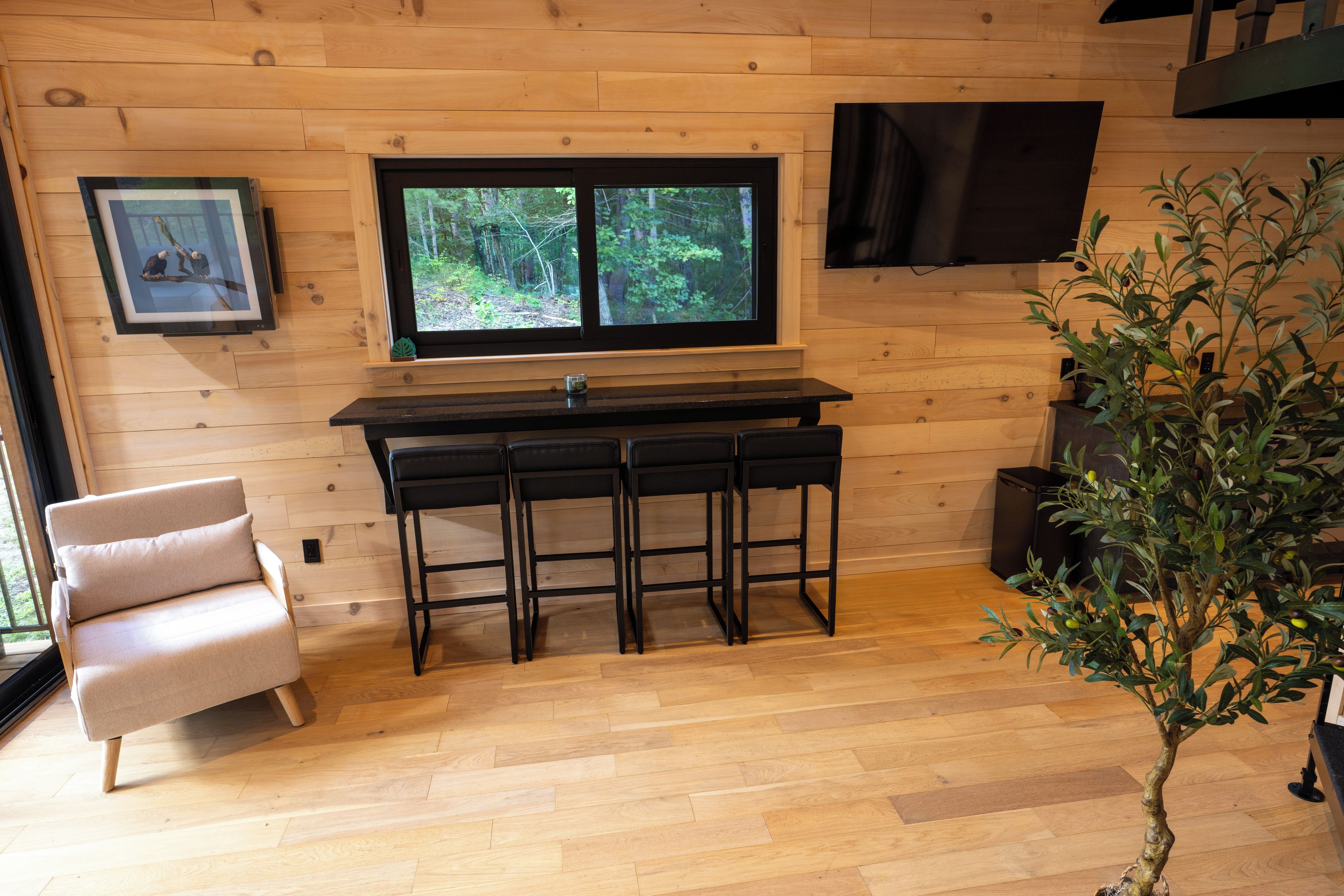 Een houten interieur met een keukenhoek, voorzien van een raam, een barkrukje, een stoel en een potplant.