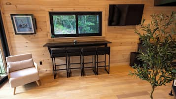Een houten interieur met een keukenhoek, voorzien van een raam, een barkrukje, een stoel en een potplant.