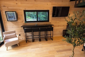 A wooden interior with a kitchen area featuring a window, a bar stool set, a chair, and a potted plant. - Glimmerwood Grove - Modern Cabin (Nassau)