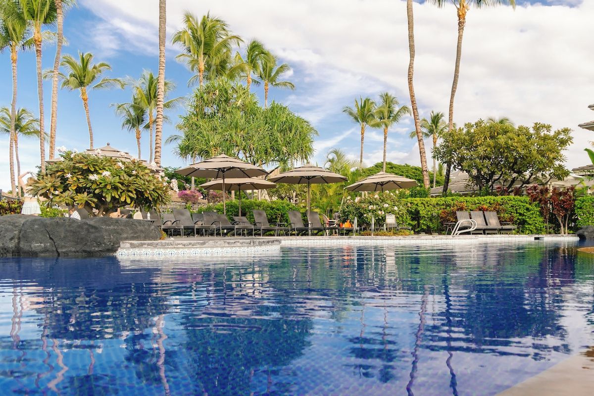 Un área de piscina con sillones de descanso y sombrillas, rodeada de palmeras y vegetación.