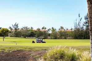 A golf course with a golf cart, trees, and buildings in the background. - Desirable Villa Walk 2 beach Light & Bright Heated Pool (Waikoloa Village)