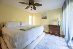 A bedroom with a large bed, a ceiling fan, a nightstand with a lamp, and a window with curtains. - Seabrook Loop by Sandpiper | Near Beach (Seabrook Island)