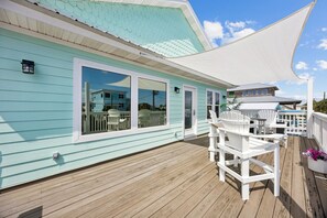 Une terrasse en bois avec des meubles d’extérieur blancs, un espace couvert avec un auvent blanc, et un bâtiment turquoise doté de grandes fenêtres.