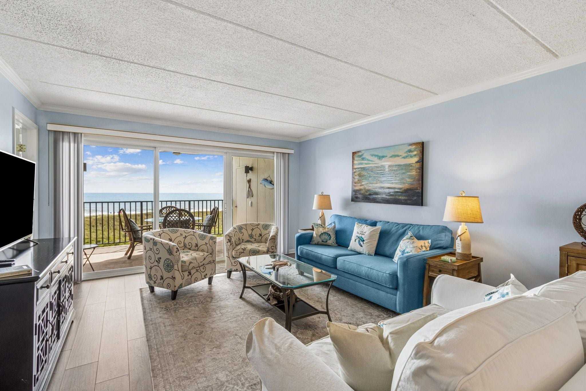 A living room with a sofa, armchairs, a coffee table, and a TV. There is a balcony with a view of the beach.