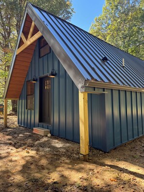 A small, single-story building with a sloped roof, a wooden front door, and a small porch area. - Charming cabin in the heart of Hocking Hills! (South Bloomingville)