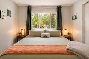 A bedroom with a large bed, two bedside tables with lamps, a window with curtains, and artwork on the walls. - Aurora Pines, a cozy Poconos retreat. (Tobyhanna)