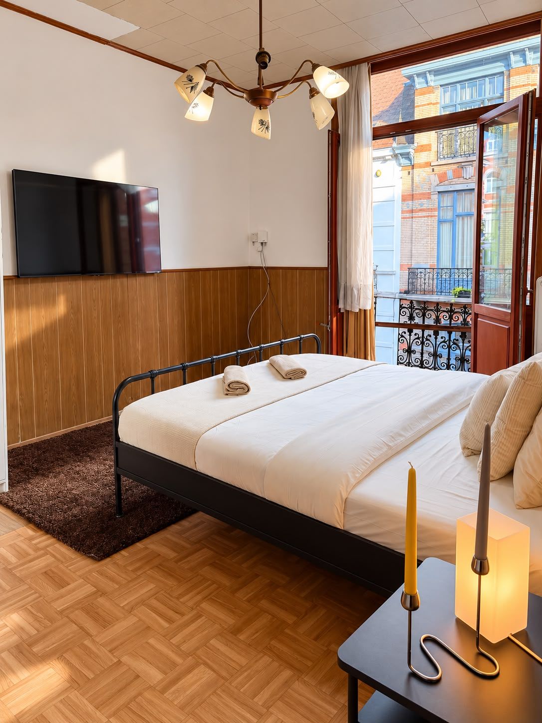 A hotel room with a bed, a flat-screen TV, a chandelier, and a window with a view of buildings.