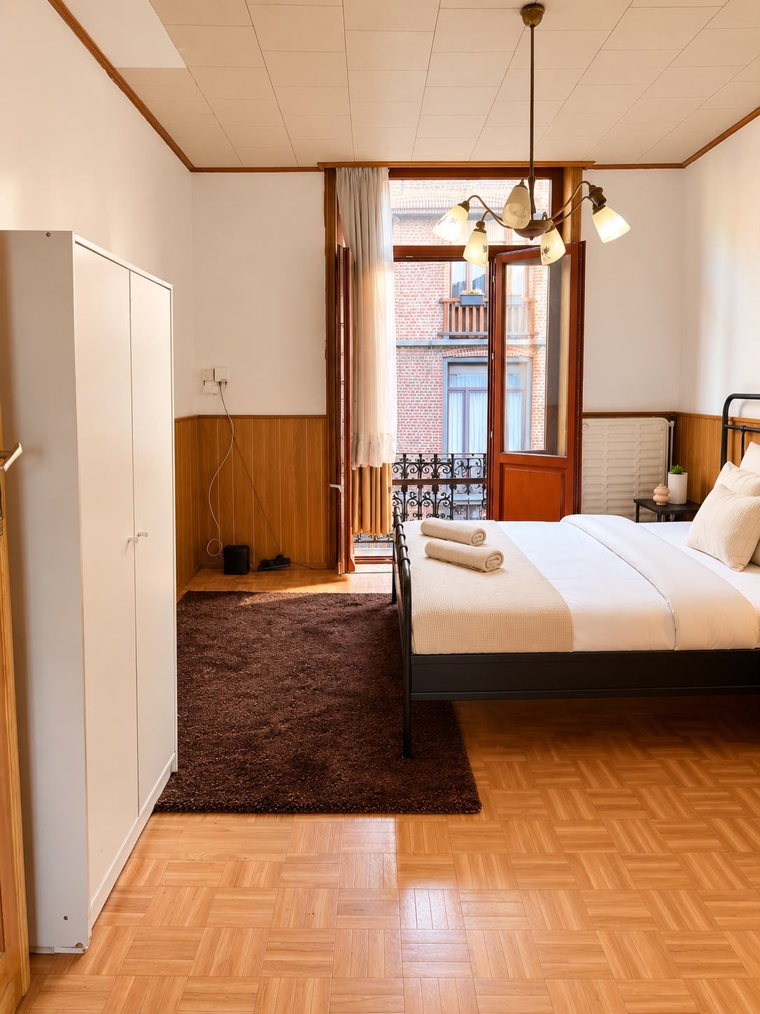 Un dormitorio con cama, armario, radiador y vista a un edificio de ladrillo a través de una ventana.