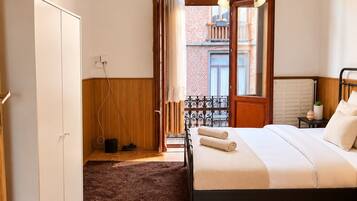 Un dormitorio con cama, armario, radiador y vista a un edificio de ladrillo a través de una ventana.