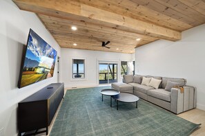 Ein modernes Wohnzimmer mit einem großen Flachbildfernseher, einem grauen Ecksofa, zwei kleinen Beistelltischen und einem grünen Teppich.