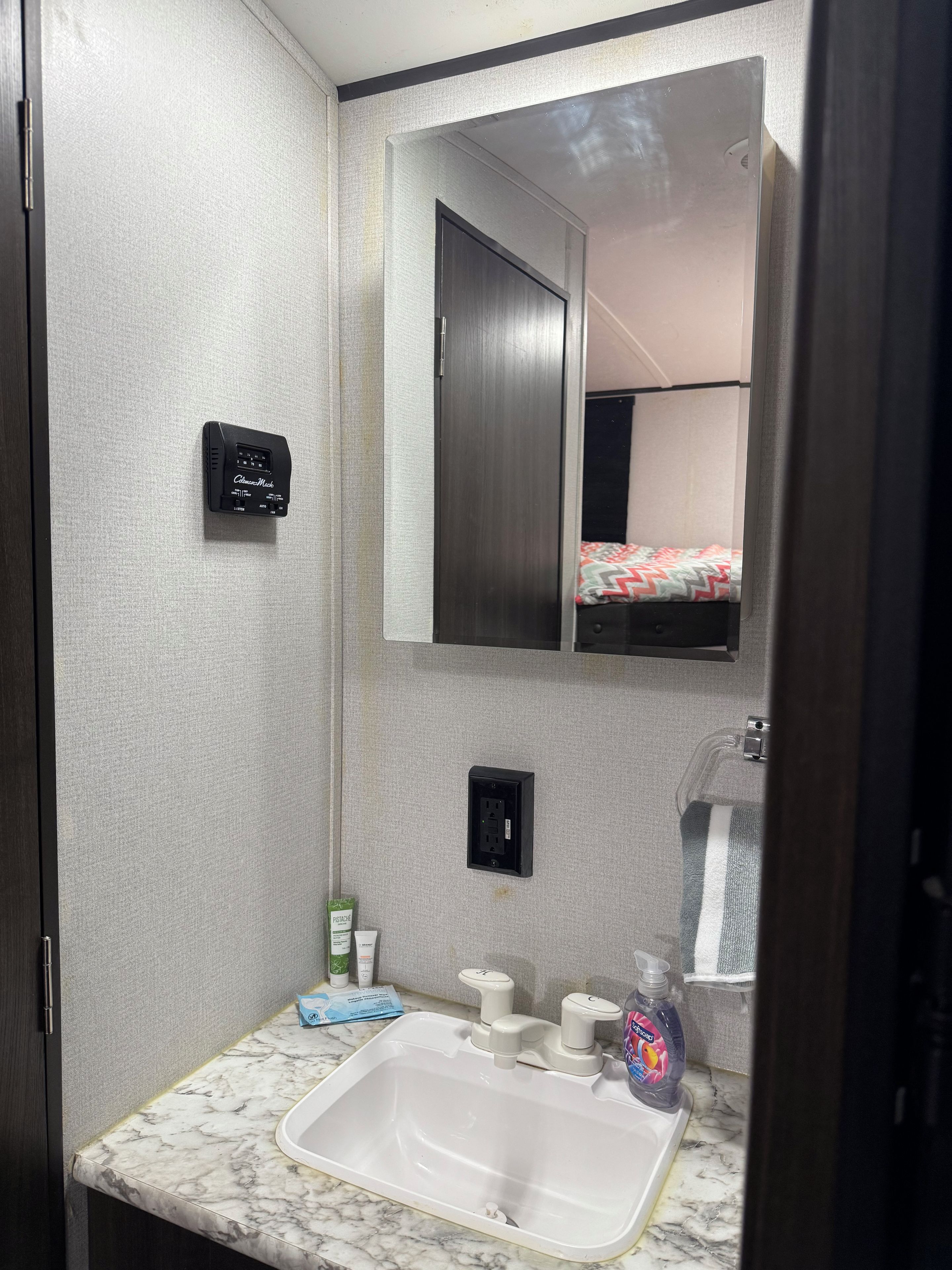Un baño con lavamanos de mármol, un espejo, un termostato y una cama visible en el reflejo.