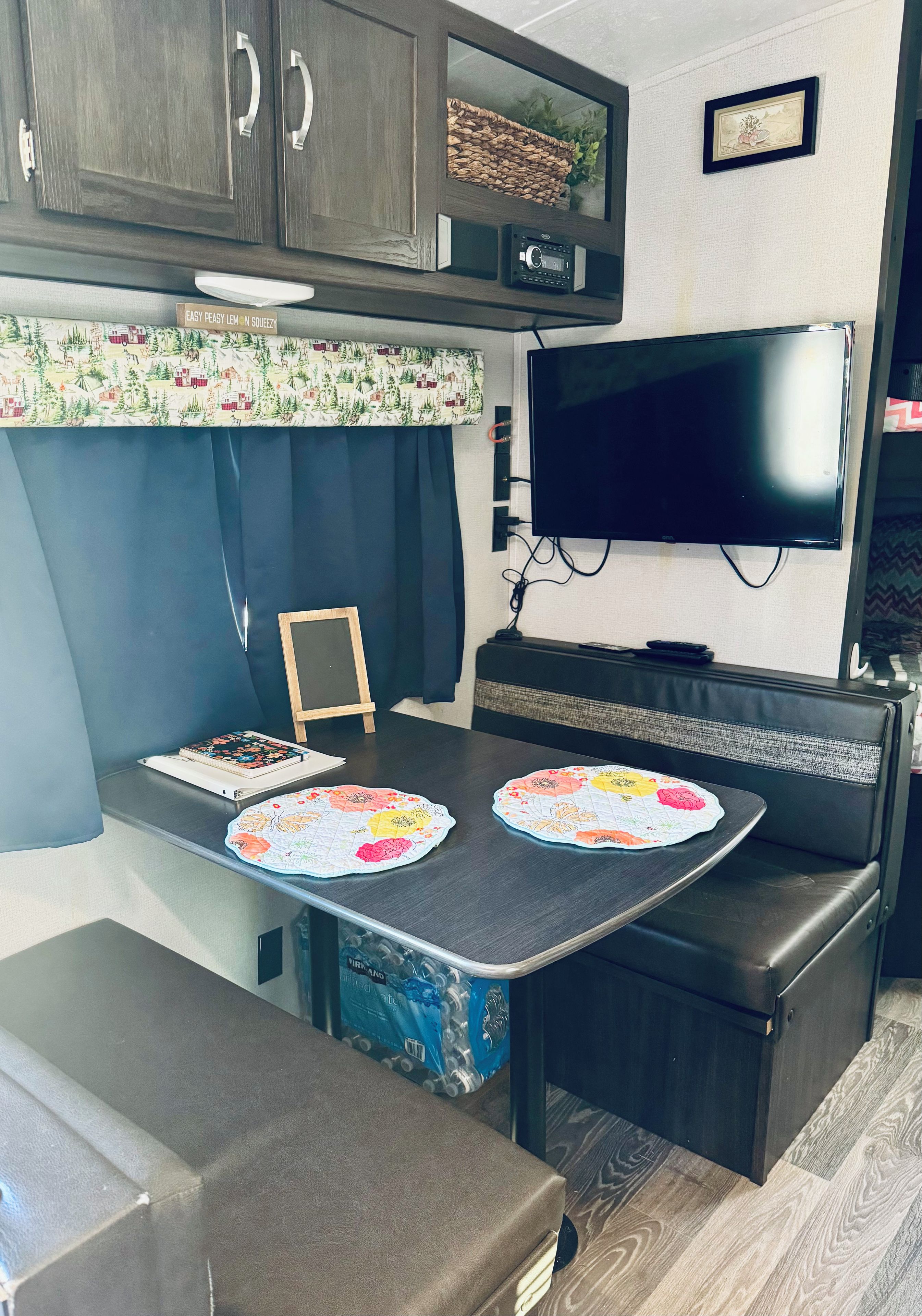 A compact RV kitchen with a flat-screen TV, a small table with a floral placemat, and a bench seating area.