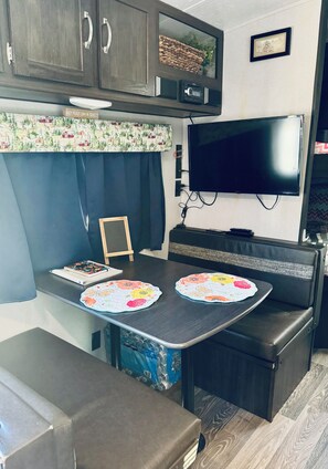 Cozinha compacta de RV com televisão de tela plana, uma pequena mesa com jogo americano floral e área de assentos com bancos.
