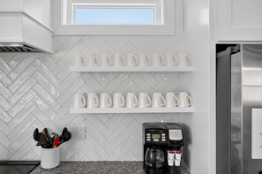 A kitchen with white herringbone patterned backsplash, two white shelves with white mugs, a coffee machine, and a utensil holder. - Coastal Paradise - Miramar Beach Escape with Pool & Golf Cart (Miramar Beach)