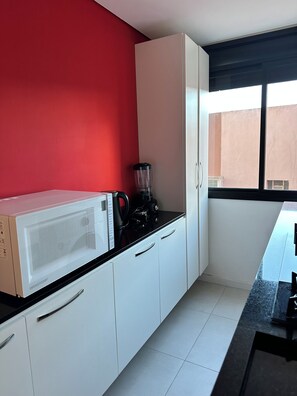 A kitchen with white cabinets, a microwave, a blender, and a window with a view of a building. - Fully furnished loft with king-size bed. (Pelotas)