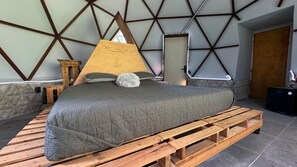 A bed with a grey quilted mattress on a wooden platform, a wooden headboard, and a wall with a geometric pattern. - Casa Libelula - Eco Glamping - Valle de Bravo (Valle de bravo)