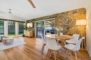 A modern dining area with a stone wall, a round table, white chairs, and a ceiling fan. - Tropical 3-Bedroom Retreat Steps to the Beach (Holloways Beach)